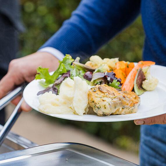 Guest holding a plate of mashed potatoes, grilled chicken, and vegetables at an outdoor buffet catering event.