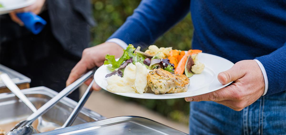Guest holding a plate of mashed potatoes, grilled chicken, and vegetables at an outdoor buffet catering event.